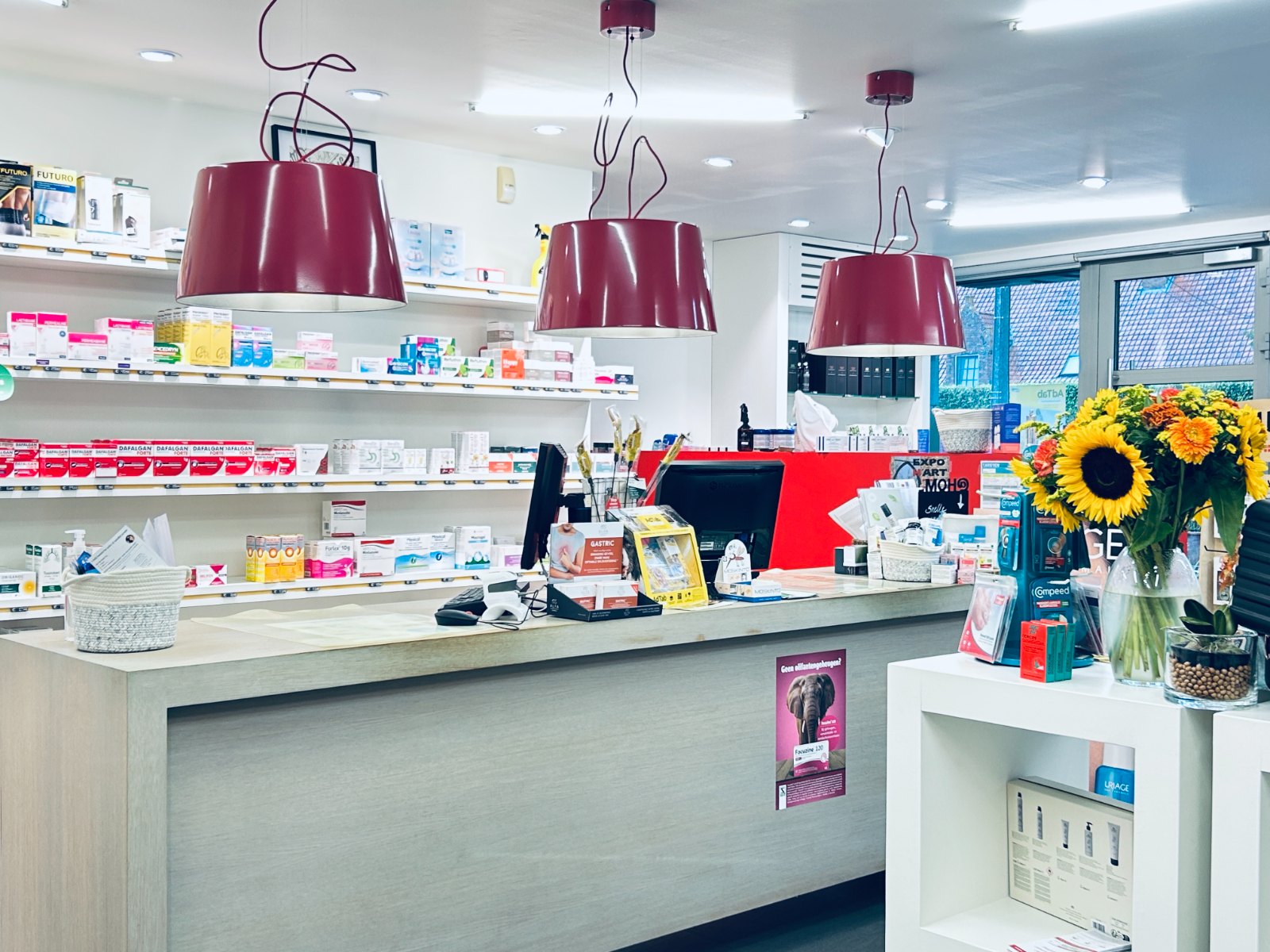 De toog van Apotheek Vansuyt met de bordeaux hanglampen en de rekken met geneesmiddelen
